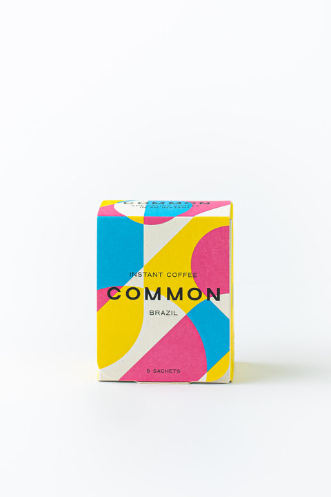 A colorful package of 'Common' brand instant coffee with the text 'BRAZIL' indicating the origin and '100% ARABICA COFFEE' on the front. The package is designed for single cup servings and has a modern, geometric pattern in shades of pink, blue, and yellow.